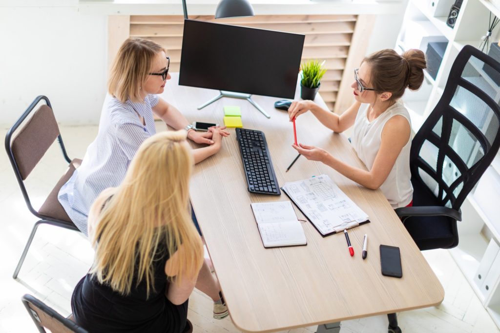 A young girl sits at a table in her office and talks to two co-partners. The girl is holding a