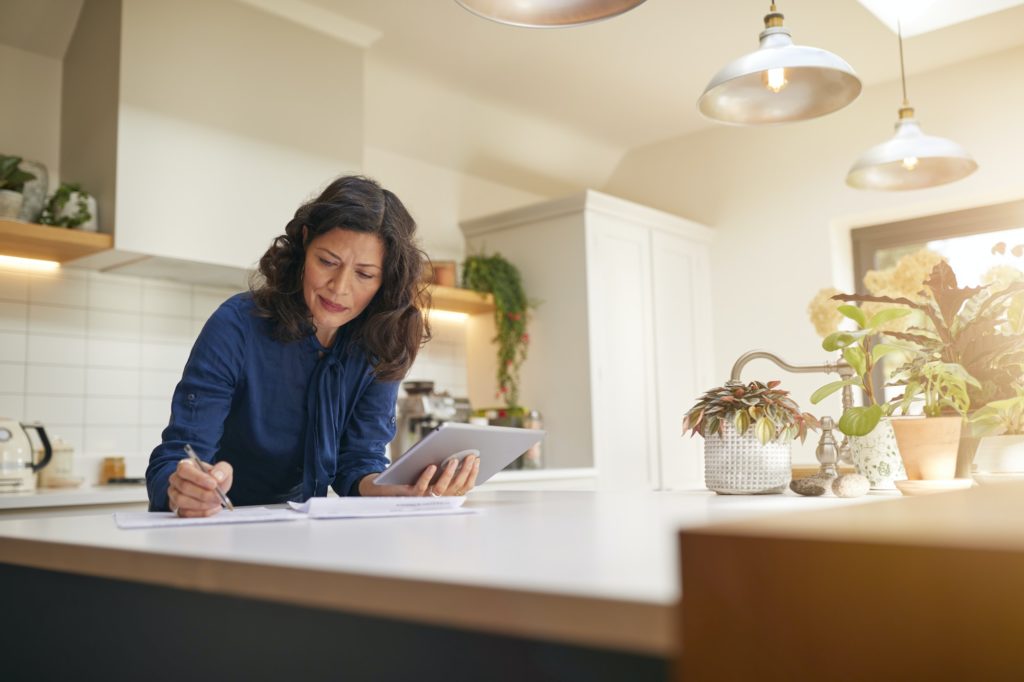 Mature Woman With Digital Tablet Reviewing Domestic Finances And Paperwork In Kitchen At Home