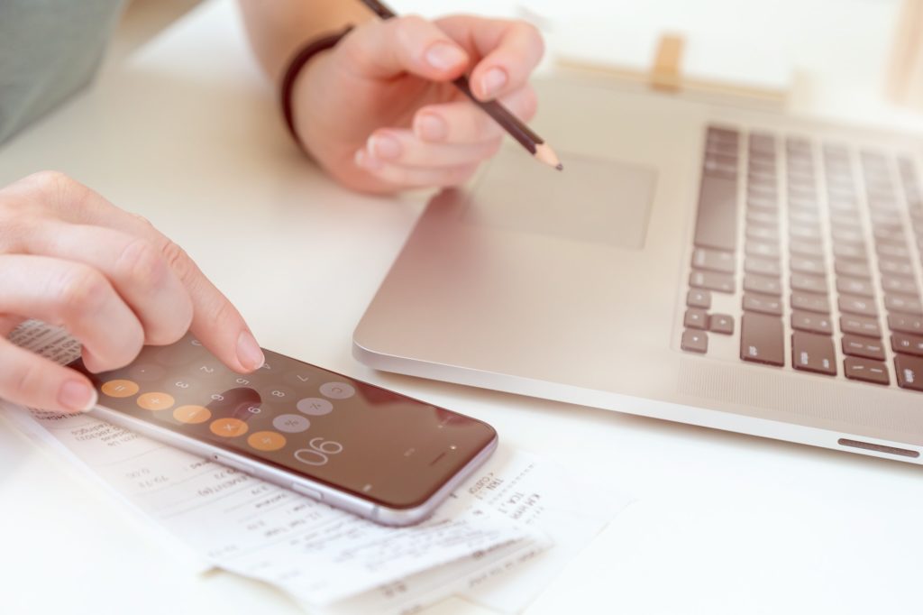 Woman making audit of household spendings at home using calculator on mobile device and laptop