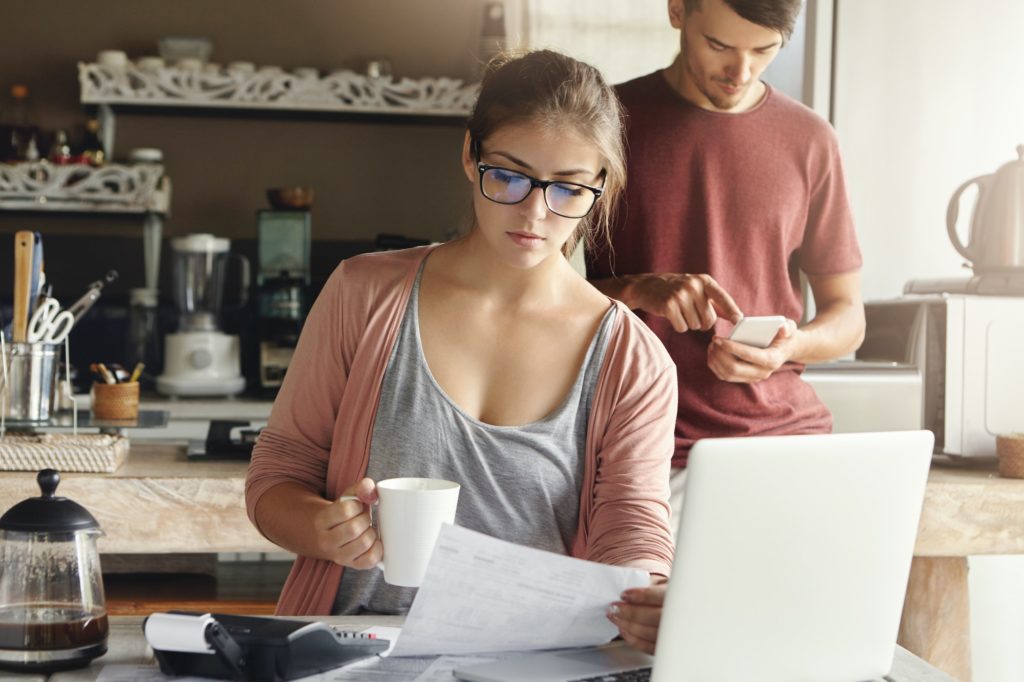 Beautiful serious young woman in rectangular glasses having coffee and studying bill in her hand, si