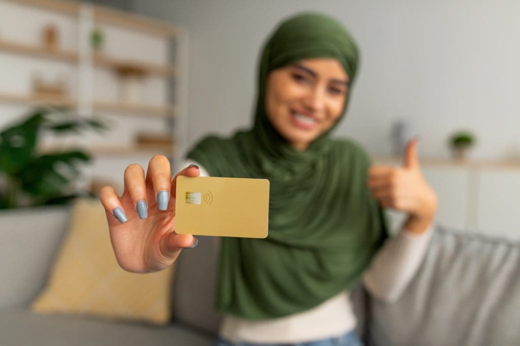 Happy young Arab woman holding credit card and showing thumb up gesture at home, selective focus